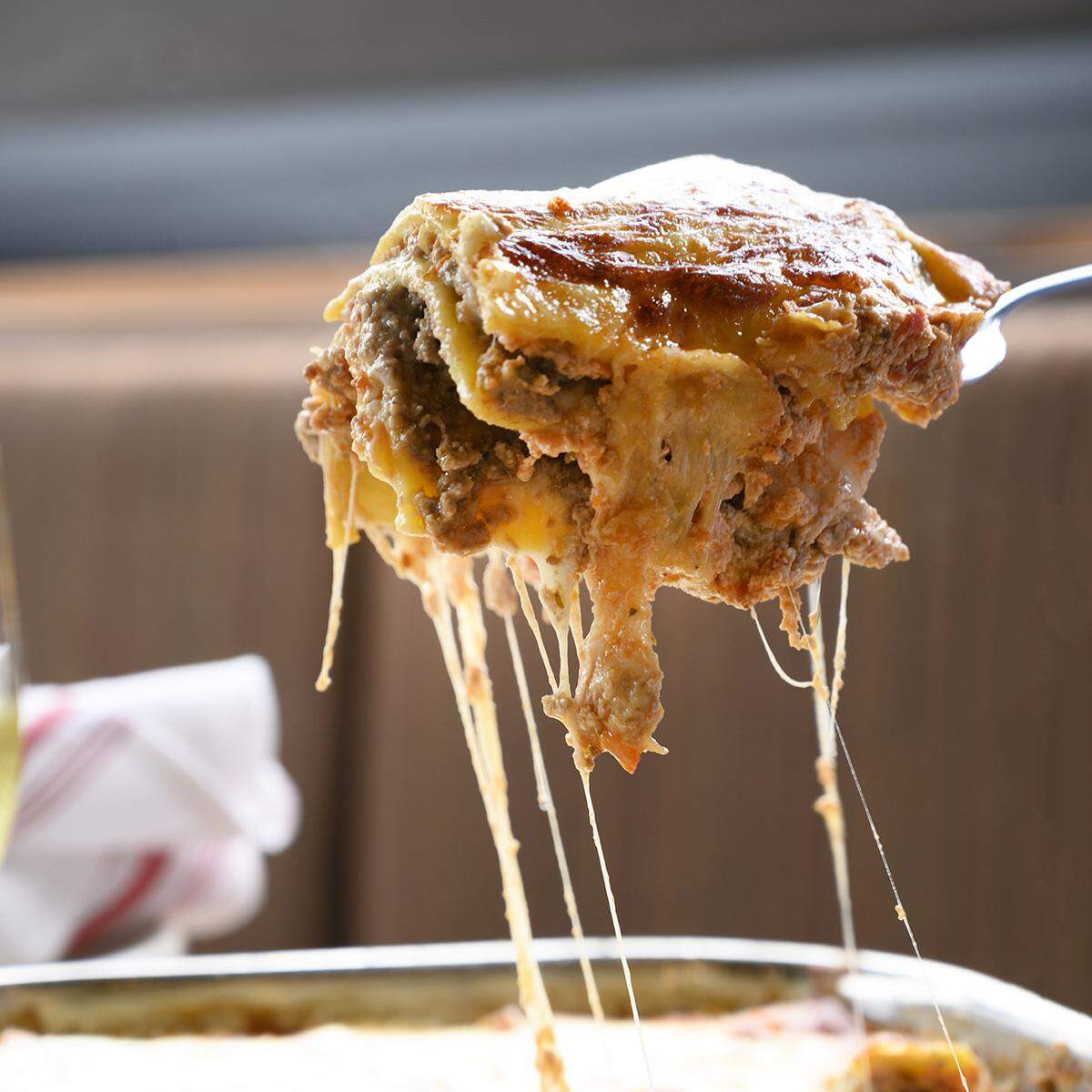 A tight, close-up shot captures a large, saucy piece of lasagna on a serving utensil being lifted, with long, dramatic strings of melted mozzarella cheese stretching down towards the pan below. The lasagna shows layers of pasta and meat sauce, with a browned cheesy top.