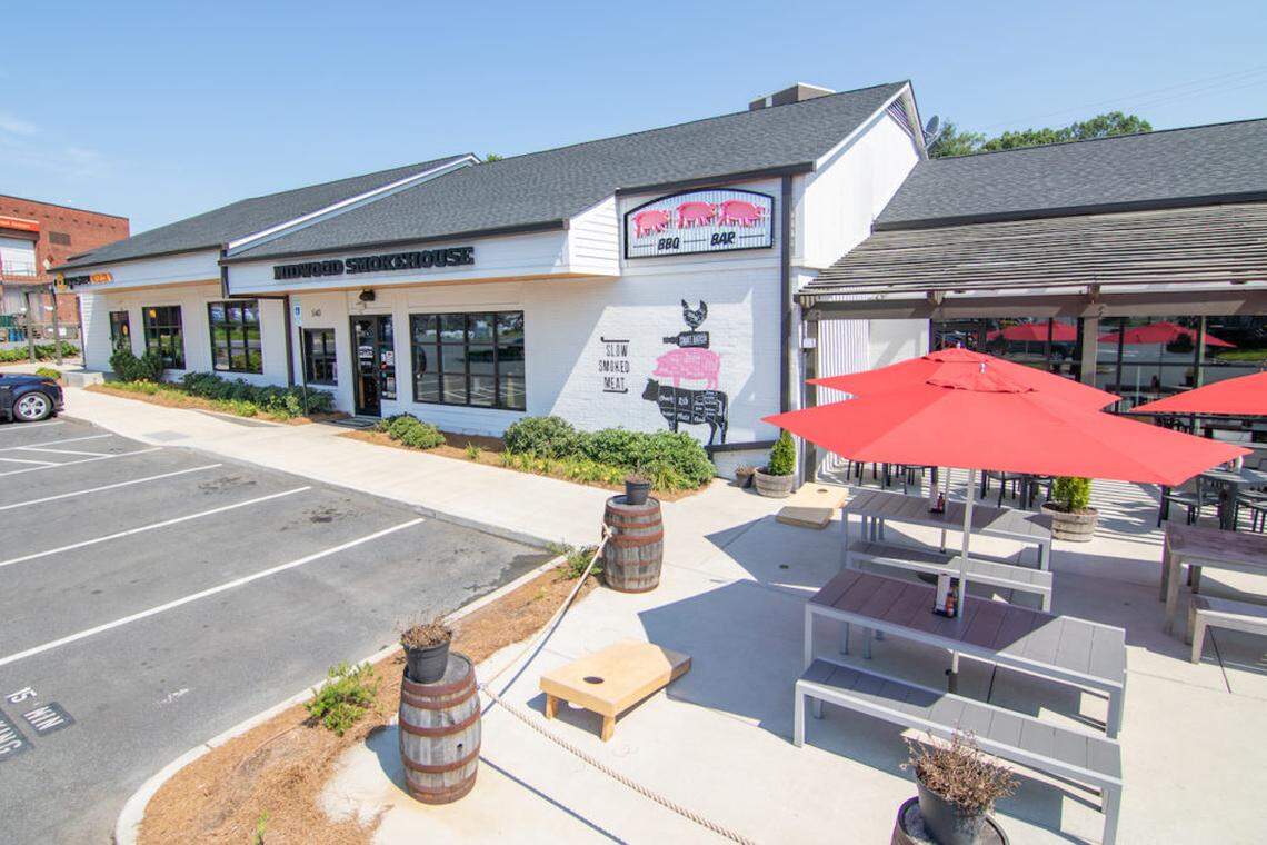  Exterior view of the Midwood Smokehouse restaurant building. The white building has large windows and an outdoor patio area with gray picnic tables and bright red umbrellas under a sunny sky.