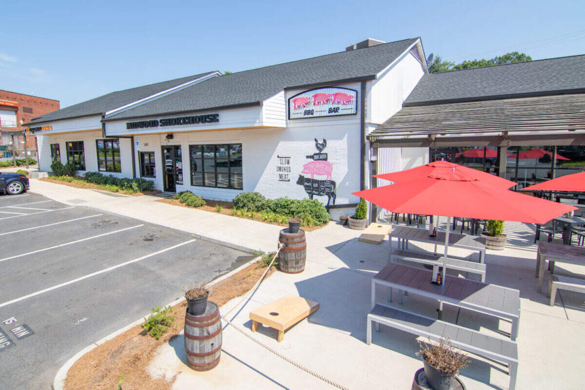 Exterior view of the Midwood Smokehouse restaurant building. The white building has large windows and an outdoor patio area with gray picnic tables and bright red umbrellas under a sunny sky.