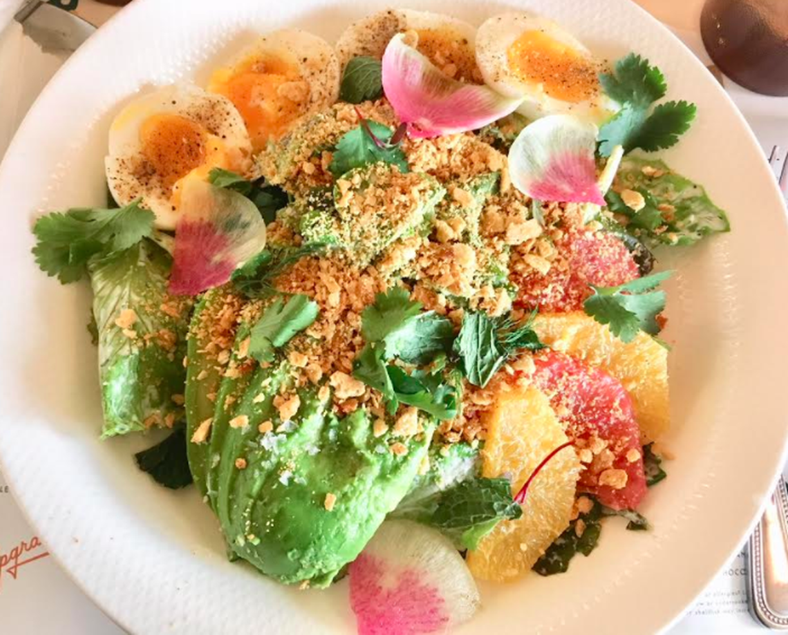 An overhead, close-up shot of a colorful Citrus Crab Louie salad in a large white bowl. The salad is artfully arranged with generous slices of avocado, halves of soft-boiled eggs, bright segments of orange and grapefruit, and fresh herbs, all topped with a generous sprinkle of a crunchy, golden-brown crumble and garnished with pink radish petals.