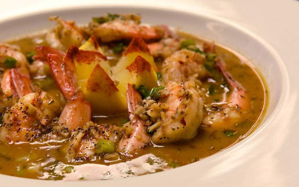 A close-up, slightly elevated shot of a bowl of New Orleans-style BBQ shrimp. Large, cooked shrimp, some with their tails still on, are generously coated in a rich, reddish-brown, buttery sauce heavily speckled with black pepper and green herbs (likely scallions or parsley). Several triangular pieces of what appear to be polenta or grits, some with a reddish sprinkle, are nestled among the shrimp. The dish is presented in a wide, white ceramic bowl.