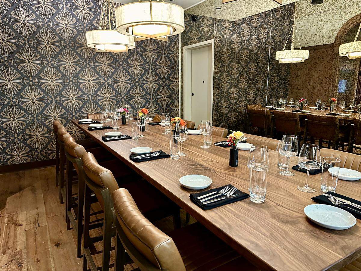A long, polished walnut dining table is set for a large group with minimalist white plates, black napkins, and sparking glassware. The room features a striking blue and gold damask wallpaper on one side and a large mirror on the other that reflects the warm glow of modern circular chandeliers. Small, colorful floral arrangements in dark vases are spaced evenly down the center of the table, adding pops of brightness to the moody, upscale space.