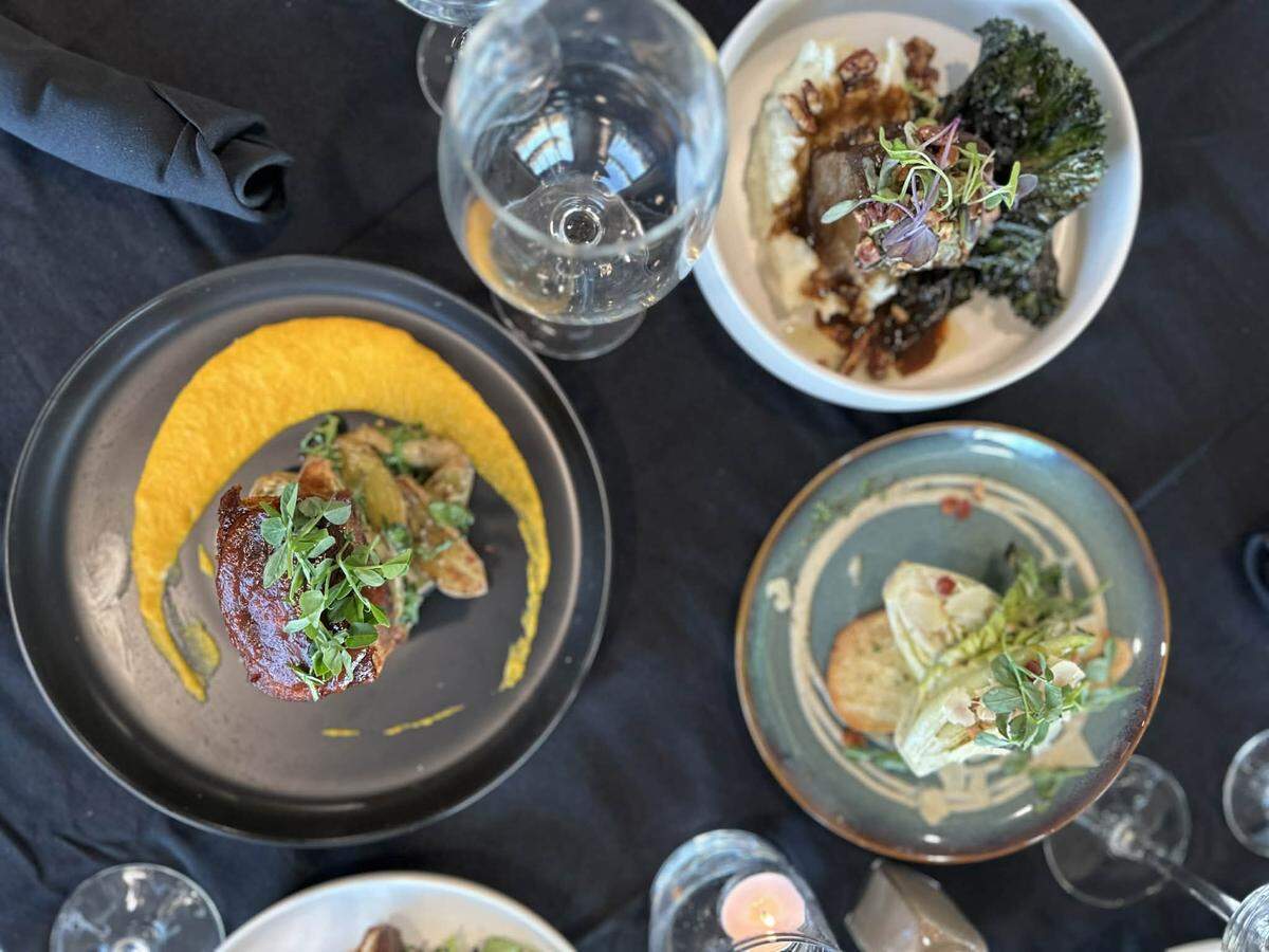An overhead shot of a dark-clothed table featuring three elegantly plated dishes, including a glazed protein with a yellow purée swoop and a side of roasted potatoes. Crystal wine glasses and a small lit candle add to the upscale, intimate dining atmosphere.
