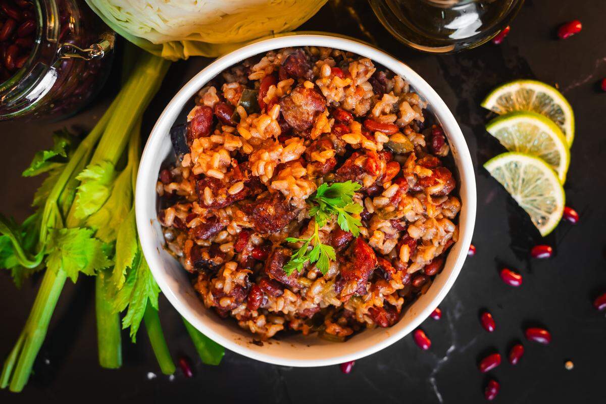 A top-down shot features a vibrant white bowl filled with Cajun red beans and rice mixed with sliced sausage and topped with a fresh parsley garnish. The bowl is staged alongside fresh celery stalks, a wedge of cabbage, and three lime wheels. Scattered loose red beans lie on the black marbled surface surrounding the plate.