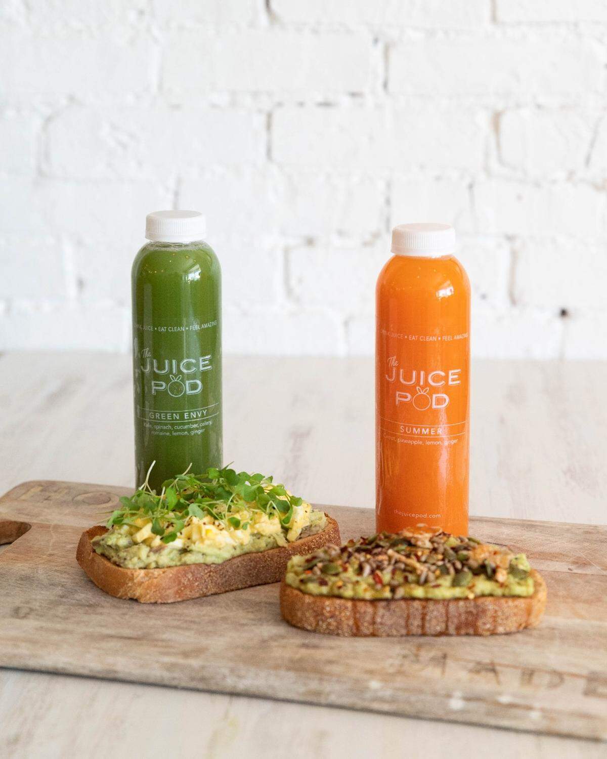 Two bottles of cold-pressed juice, one vibrant green and one bright orange, stand behind two thick slices of artisanal avocado toast. The toasts are heavily garnished with microgreens, seeds, and red pepper flakes, all presented on a wooden board against a white brick background.