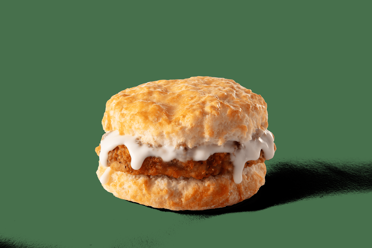 A golden-brown biscuit is sliced and holds a filling of a breaded patty, likely country fried steak, which is generously topped with white gravy. The gravy drips down the sides, contrasting with the rich color of the biscuit and steak, all set against a black background.