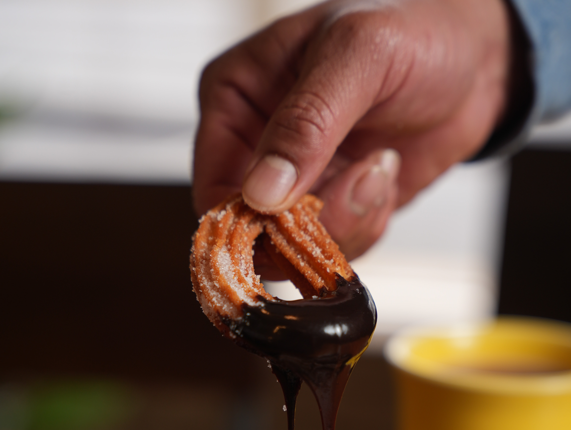 A hand holds a single, ring-shaped churro coated in sugar, which is being dipped into a pool of dark, melted chocolate. The chocolate drips from the churro, and a bright yellow cup containing more chocolate dipping sauce is partially visible in the blurred background.
