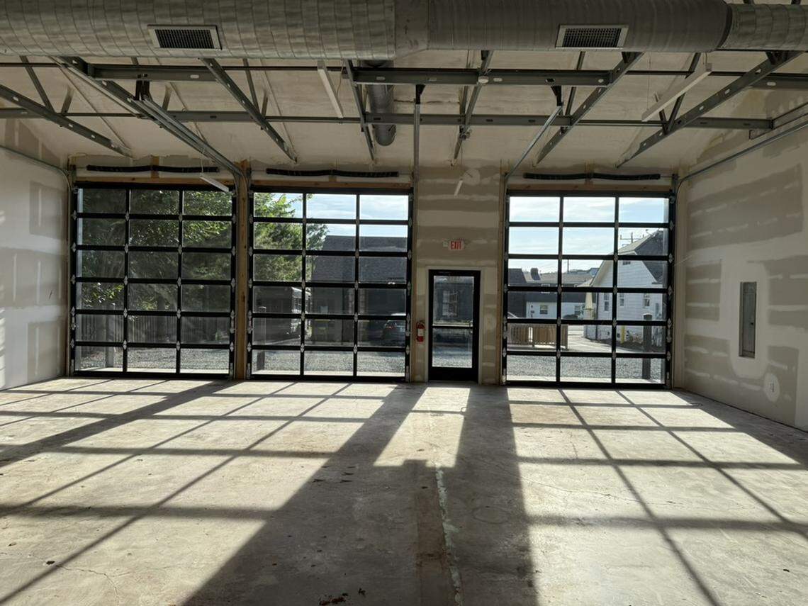 The interior of an unfinished industrial space featuring three large glass garage doors and a central glass entry door. Sunlight streams through the panes, casting long, geometric shadows across a raw concrete floor.