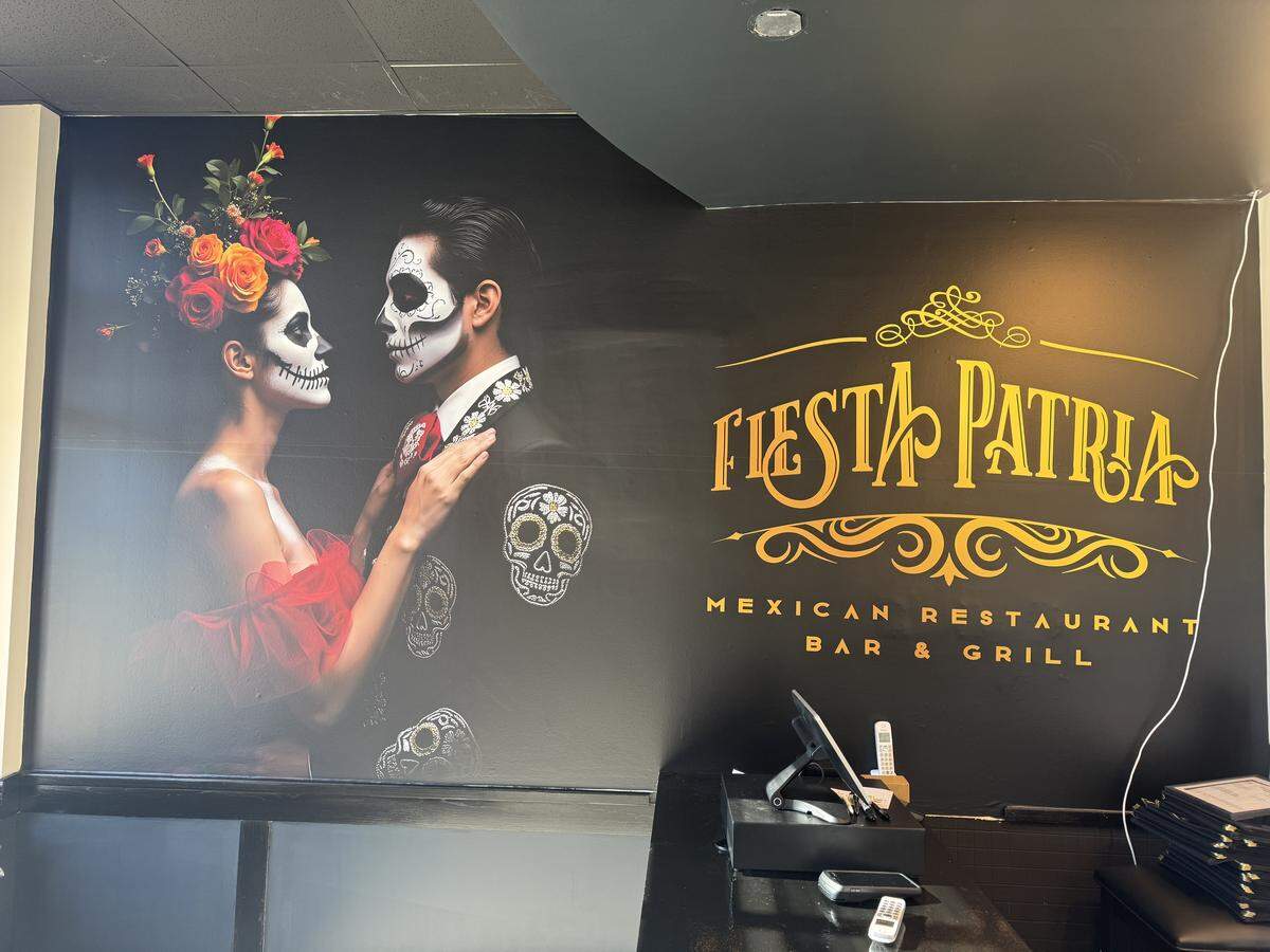 A black wall in the restaurant’s lobby features a large, colorful mural of a couple dressed with Día de Muertos or Calaca makeup and floral headpieces, facing each other lovingly. To the right of the artwork, the restaurant’s name, “FIESTA PATRIA MEXICAN RESTAURANT BAR & GRILL,” is prominently displayed in gold script.