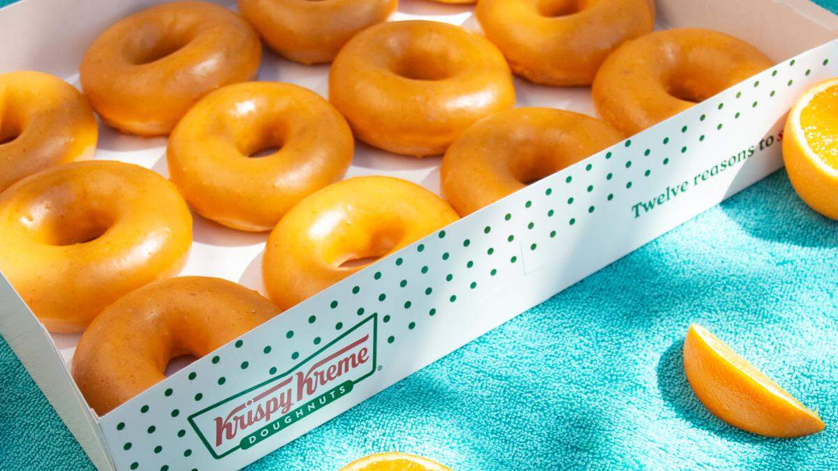 A white Krispy Kreme box filled with twelve orange-glazed donuts sits on a bright teal textured surface, likely a beach towel. Fresh orange slices are scattered around the box, and the scene is illuminated by bright, sunny outdoor light that creates soft shadows. The side of the box is visible, featuring the brand logo and the phrase “Twelve reasons to smile.”