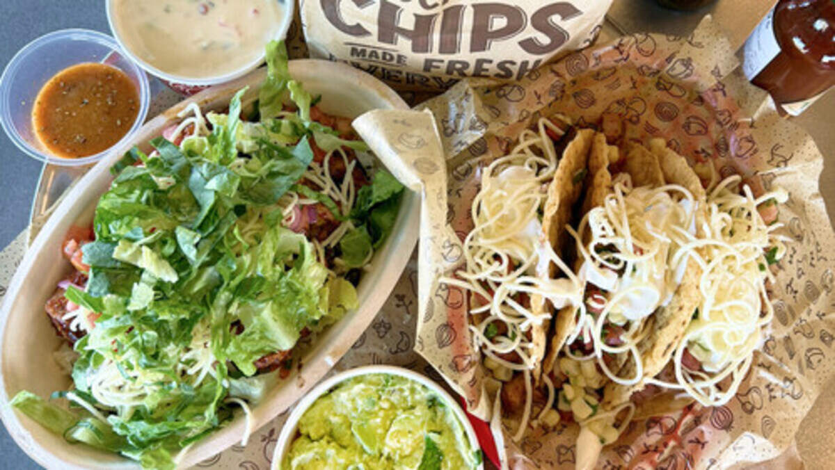 A vibrant overhead shot features a spread of Mexican-style food including a large burrito bowl topped with shredded lettuce and a basket of three tacos overflowing with cheese. The meal is accompanied by sides of guacamole, queso, salsa, and a bag of tortilla chips.