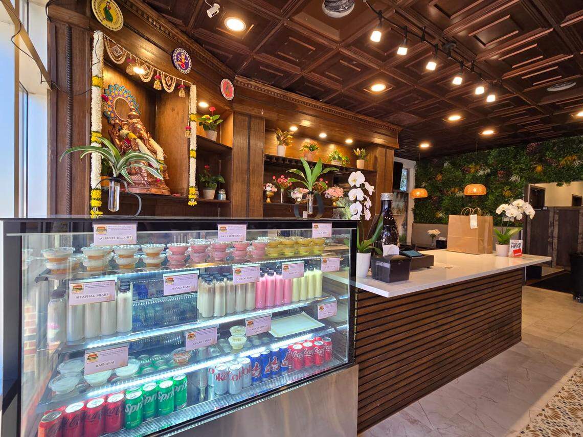 A brightly lit counter features a display case stocked with various colorful desserts and bottled drinks like flavored milk and sodas. Behind the counter, there’s a wooden shrine with a decorated statue of Ganesha and small vases of flowers.