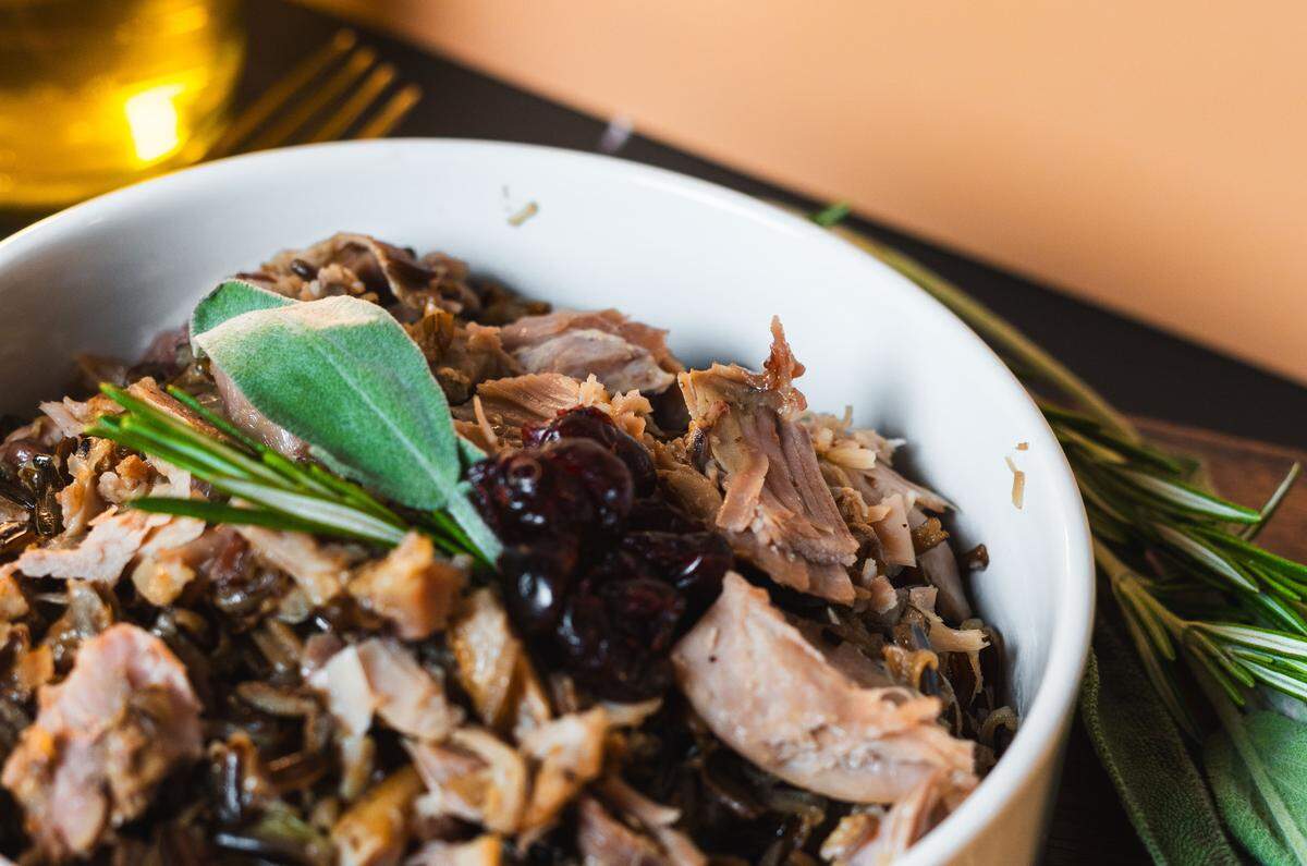 An angled shot captures a white bowl filled with shredded turkey meat mixed with wild rice. The dish is garnished with a single green sage leaf, a sprig of rosemary, and a small cluster of dark cranberries in the center. Fresh sprigs of thyme rest beside the bowl on a wooden cutting board against a warm, peach-colored background.