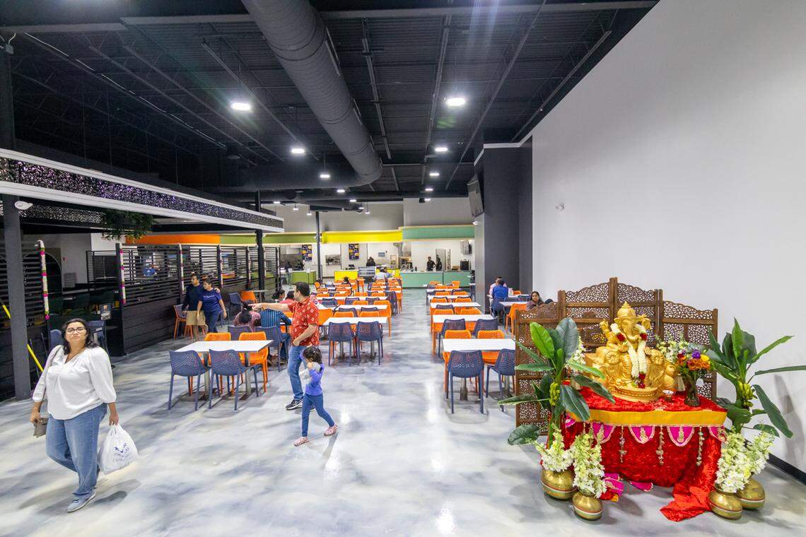 This interior photo captures the large food court dining area with a black industrial ceiling and a marbled epoxy floor. In the foreground, there is a golden Ganesha statue on a red altar, while customers are seated at tables or walking through the space.