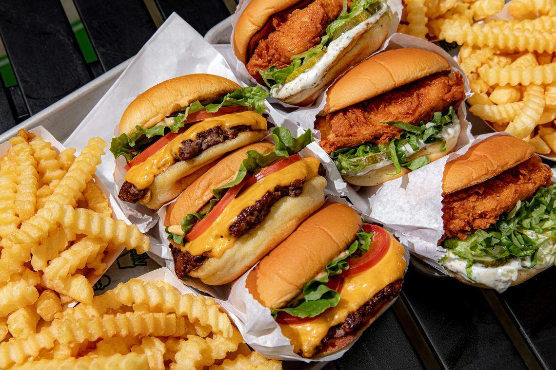 A high-angle, close-up shot of a metal tray packed with three cheeseburgers, three crispy chicken sandwiches, and two generous portions of crinkle-cut fries. The sandwiches are neatly tucked into white paper sleeves, showcasing fresh lettuce, tomato, and melted cheese.