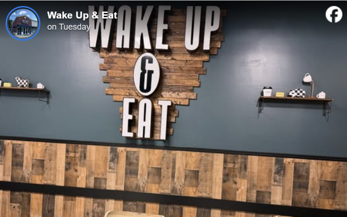 A large, stylized wall sign reads “WAKE UP & EAT” in bold block letters, with the words mounted on an inverted triangle of rustic wood planks against a dark teal wall. Below the teal wall is a lower section of paneling made from multi-toned, vertical wooden boards. Small decorative shelves are mounted on either side of the main sign, displaying tiny knick-knacks like a coffee cup and a patterned pig figurine.