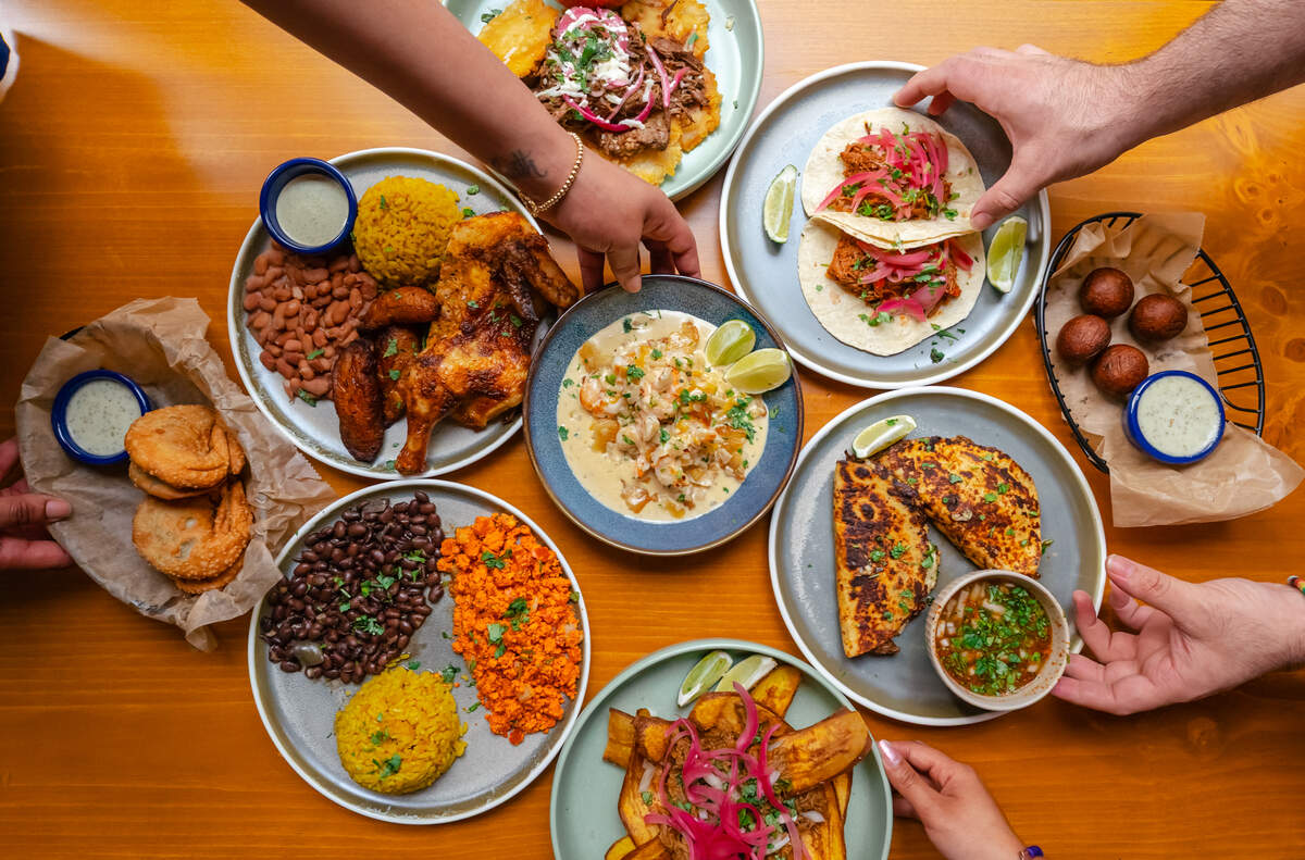 A top-down, flat-lay photograph captures a vibrant feast on a wooden table as multiple hands reach in to share the dishes. The spread includes various plates of yellow rice, black beans, sweet plantatins, roasted chicken, soft tacos, and crispy empanadas. In the center, a creamy shrimp dish serves as the focal point, surrounded by small bowls of dipping sauces and fresh garnishes.