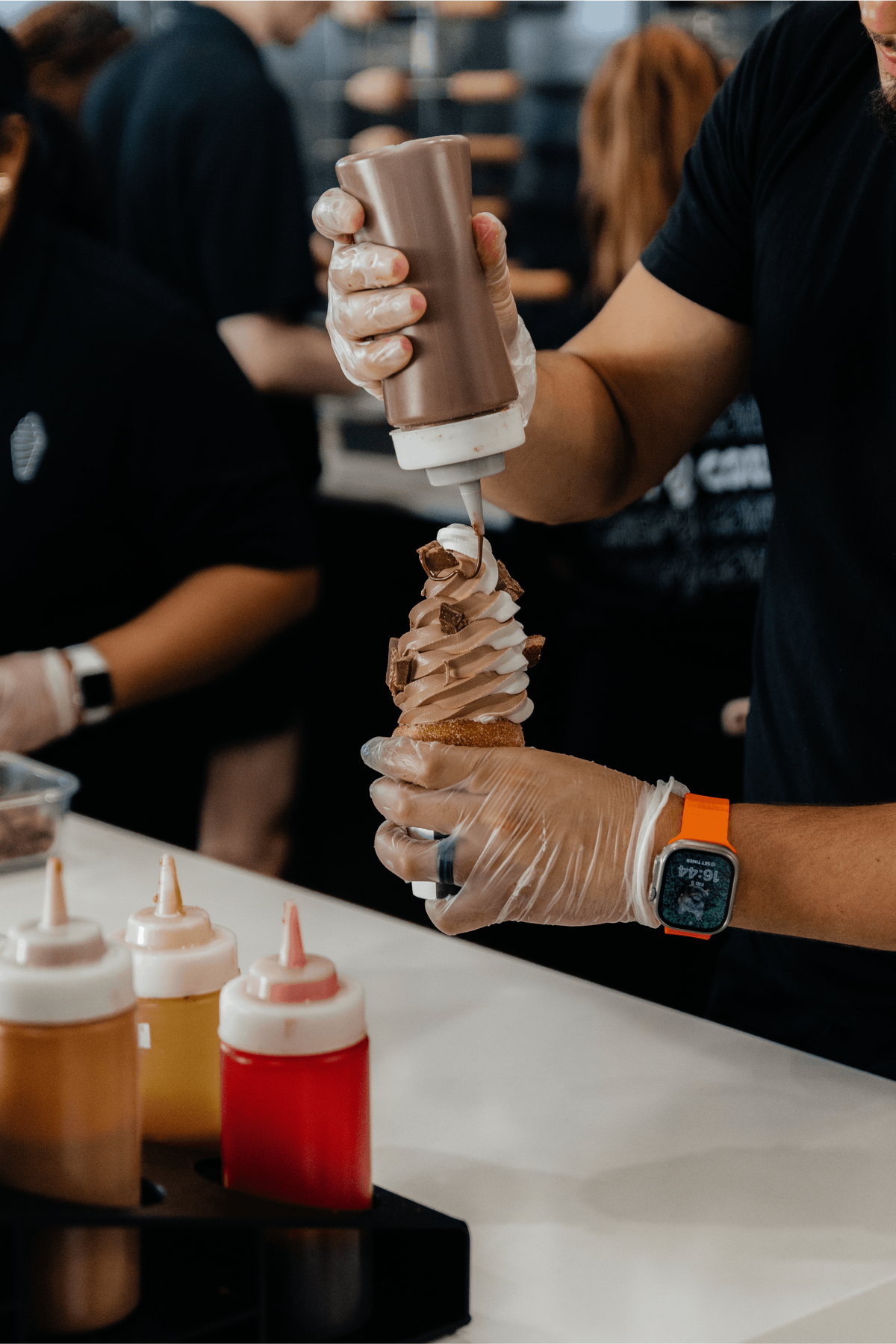 A worker wearing clear plastic gloves uses a squeeze bottle to drizzle chocolate sauce over a tall swirl of chocolate and vanilla twist soft-serve ice cream. The ice cream is piled high inside a pastry cone, garnished with chocolate chunks, with several squeeze bottles lined up on the counter in the foreground.