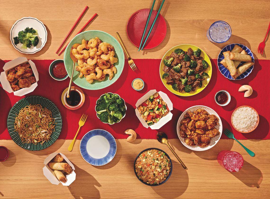 An overhead spread of American Chinese dishes including orange chicken, honey walnut shrimp, and chow mein arranged on a wooden table with a red runner. Colorful plates, bowls, and takeout containers are scattered throughout the scene alongside chopsticks and fortune cookies.