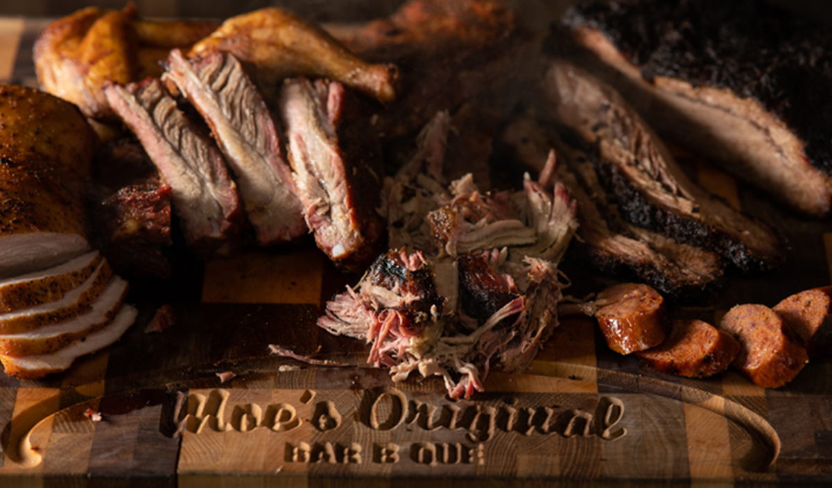 A large wooden cutting board, engraved with “Moe’s Original BBQ,” displays a smoky assortment of sliced turkey, ribs, pulled pork, and smoked chicken. Thick slices of spicy sausage and a dark, charred brisket rest on the right, showcasing a variety of textures and traditional pit-cooking techniques. The dramatic, low-key lighting emphasizes the glistening juices and blackened bark of the various meats.