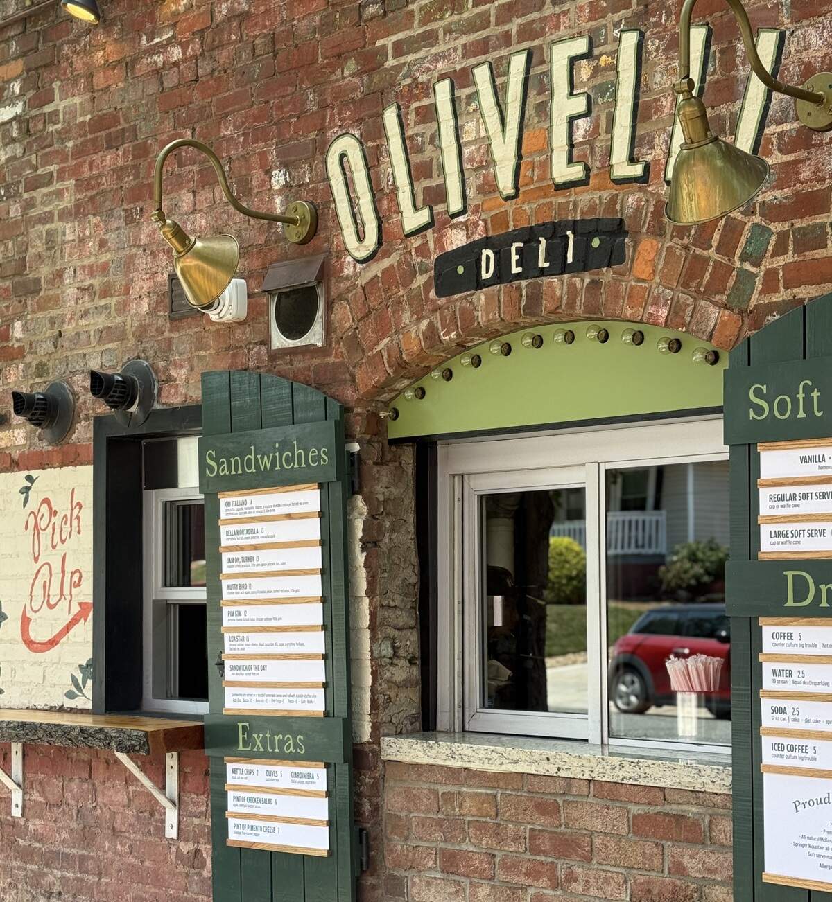 A close-up shot shows the brick exterior of Olivelli Deli, featuring a walk-up ordering window and a wooden “Pick Up” sign to the left. Hanging green shutters display the menu, which includes sections for sandwiches, extras, soft serve, and drinks. Two brass gooseneck lamps illuminate the scene from above the arched window frame.