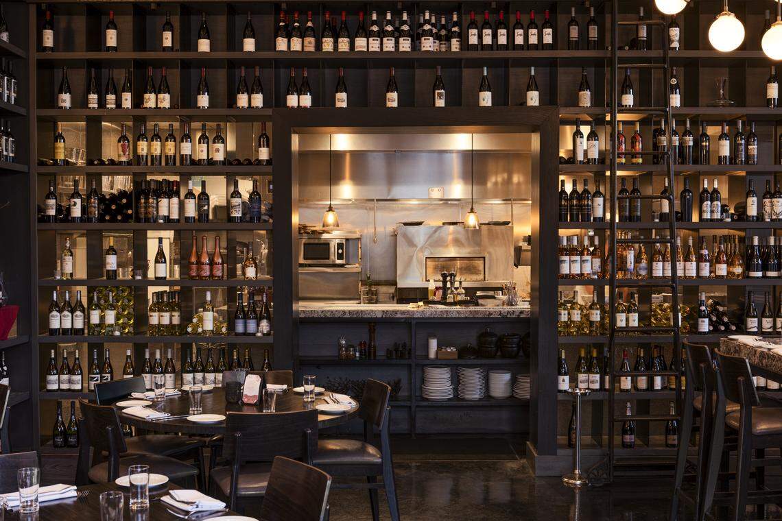 A wide shot of the restaurant interior showcases a massive, floor-to-ceiling wine wall filled with hundreds of bottles, surrounding a central opening that looks into a clean, stainless steel kitchen. In the foreground, neatly set circular dining tables with dark chairs are ready for guests under warm, ambient lighting.