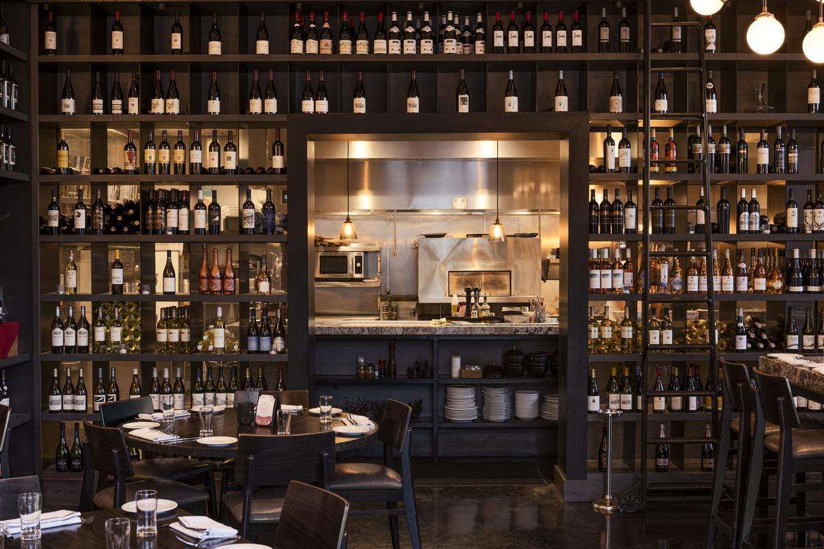 A wide shot of the restaurant interior showcases a massive, floor-to-ceiling wine wall filled with hundreds of bottles, surrounding a central opening that looks into a clean, stainless steel kitchen. In the foreground, neatly set circular dining tables with dark chairs are ready for guests under warm, ambient lighting.