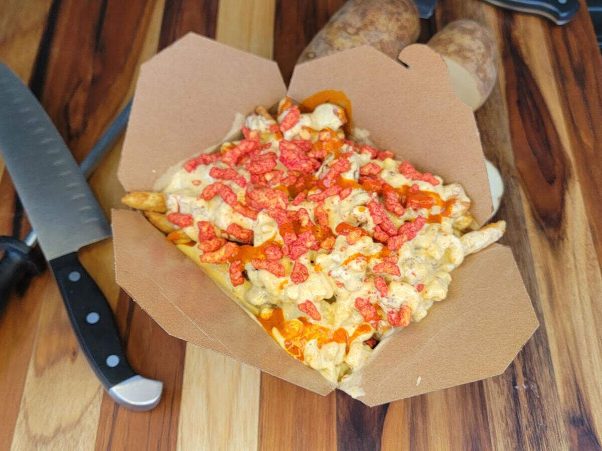 A unique and colorful dish of loaded fries is presented in a cardboard container, featuring fries smothered in a creamy white cheese sauce and a vibrant drizzle of bright red hot sauce. The entire creation is then generously topped with bright red, crumbled Flamin’ Hot Cheetos, positioned on a wooden surface next to raw potatoes and a knife.
