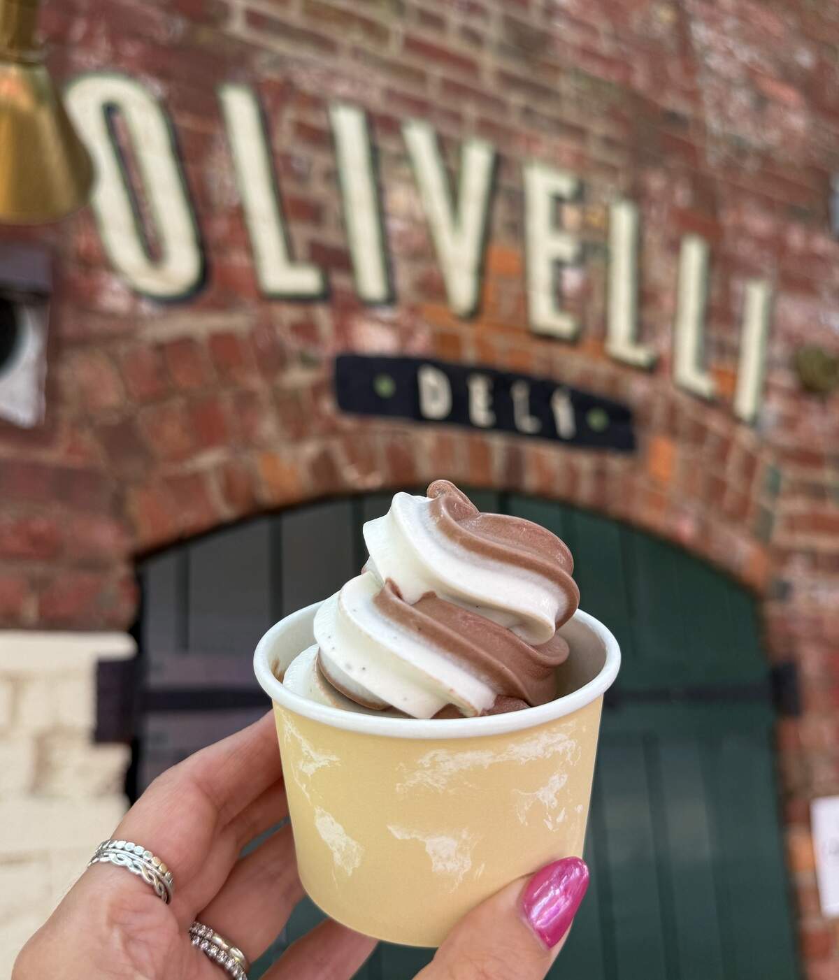 A hand with pink nail polish and silver rings holds up a small paper cup filled with chocolate and vanilla twist soft serve ice cream. In the softly blurred background, the brick facade and painted “Olivelli Deli” sign are visible. The treat is positioned centrally, showcasing the clean swirl of the dessert.