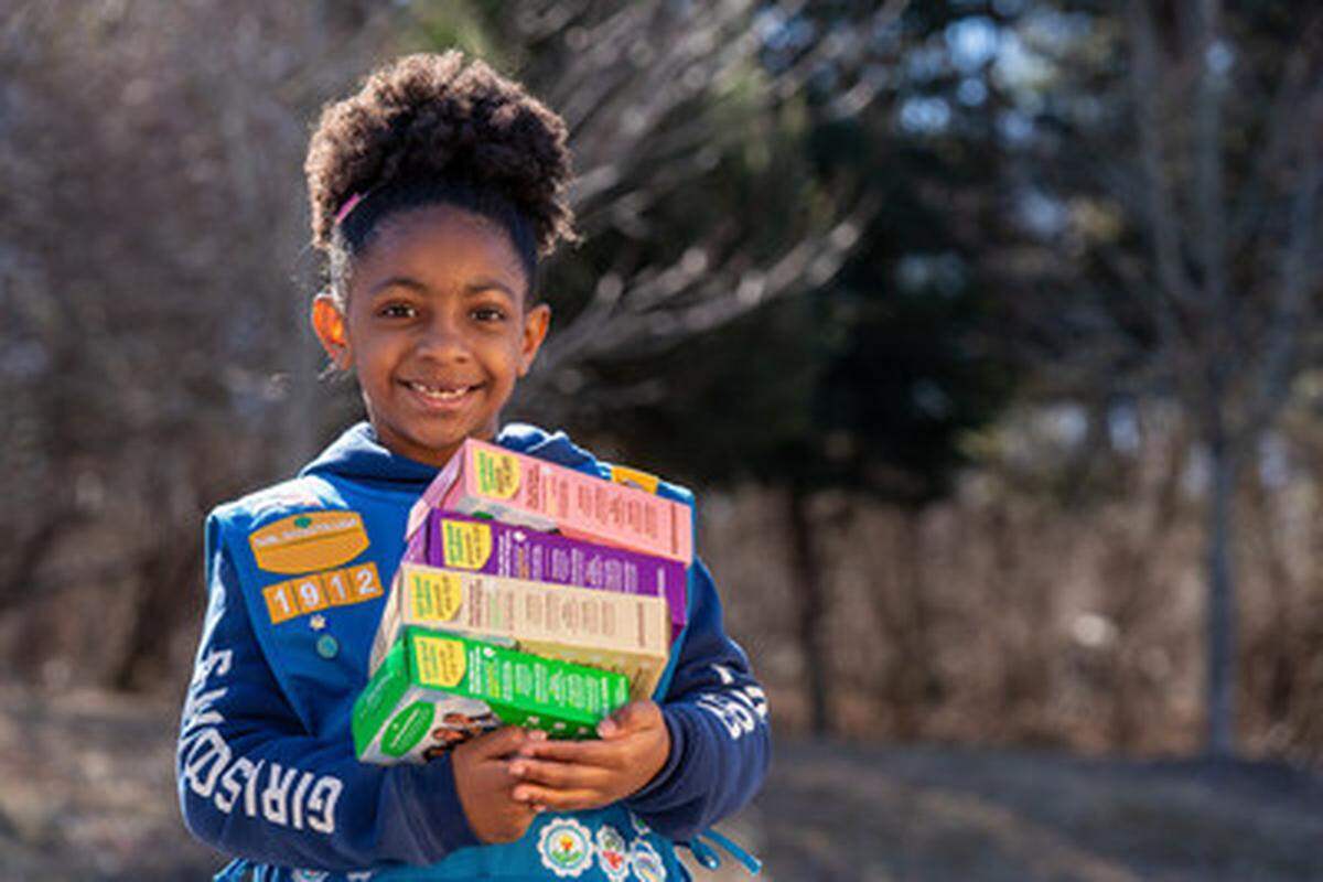 A smiling Girl Scout in a blue vest stands outside while balancing a large stack of various cookie boxes in her arms. The background features a soft-focus wooded area, highlighting the scout and her colorful collection of cookie packages.