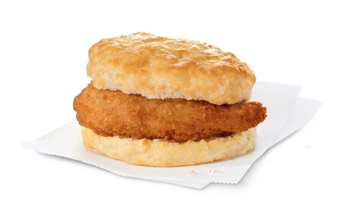 A breaded, golden-brown chicken breast fillet is served on a warm, flaky buttermilk biscuit. The breakfast sandwich rests on a white parchment paper against a clean, solid white background.