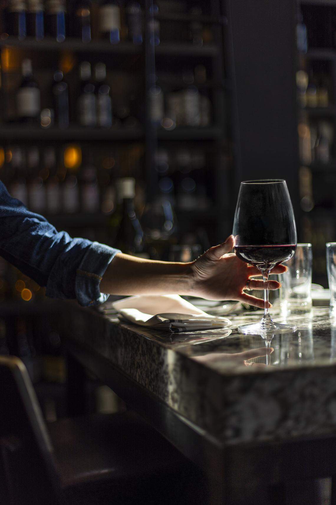 A close-up, atmospheric shot shows a person’s arm reaching toward a large glass of red wine resting on a polished marble bar top. The background is dimly lit, featuring out-of-focus shelves filled with numerous wine bottles, creating a cozy and upscale mood.