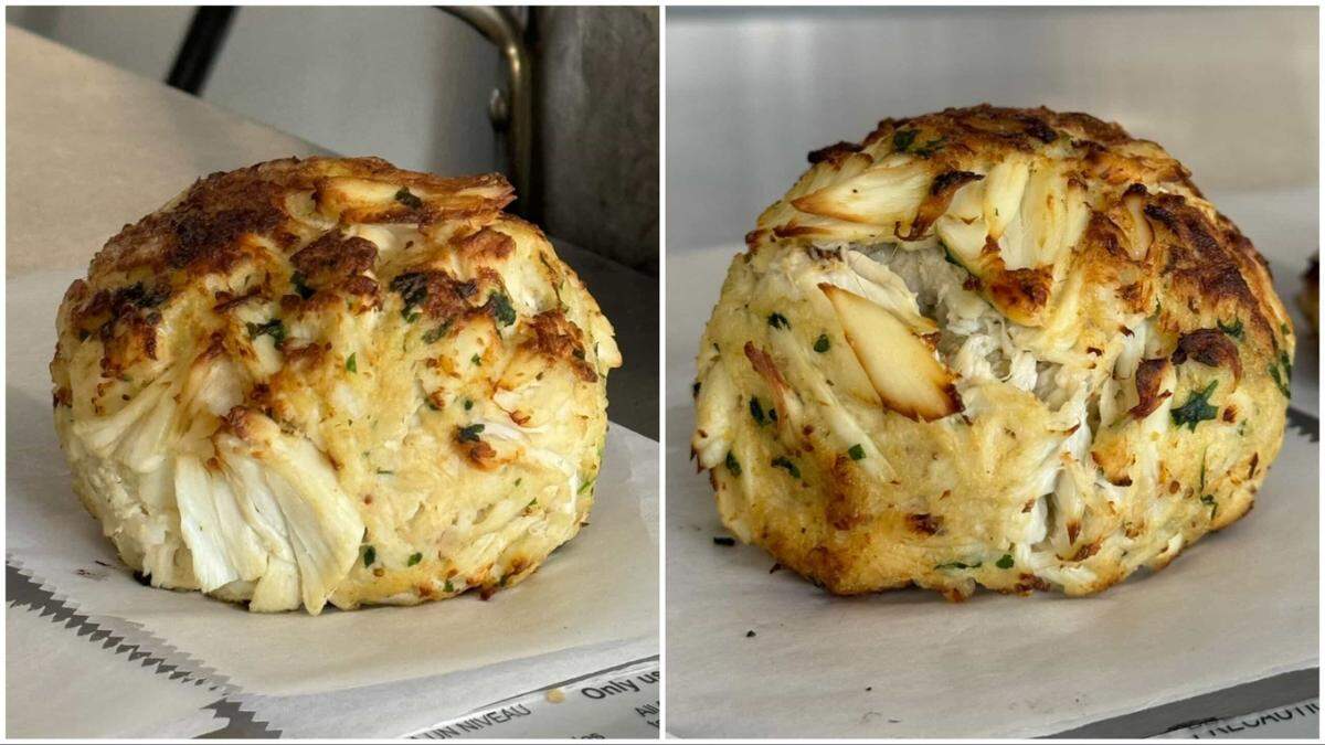 A large, spherical crab cake packed with colossal lumps of white crab meat and flecks of fresh green herbs sits on white parchment paper. The top is golden-brown and toasted, showcasing the dense, premium texture of the seafood.