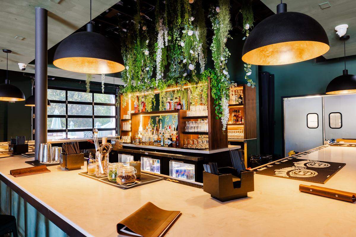 A close-up view looks across the smooth, light-colored surface of the bar counter, showcasing leather-bound menus, cocktail strainers, and jars of garnishes. The central liquor island is brightly backlit, highlighting various bottles, glassware, and refrigerated beverage coolers below. Lush faux vines and white floral arrangements hang directly over the center structure, creating a vibrant canopy.
