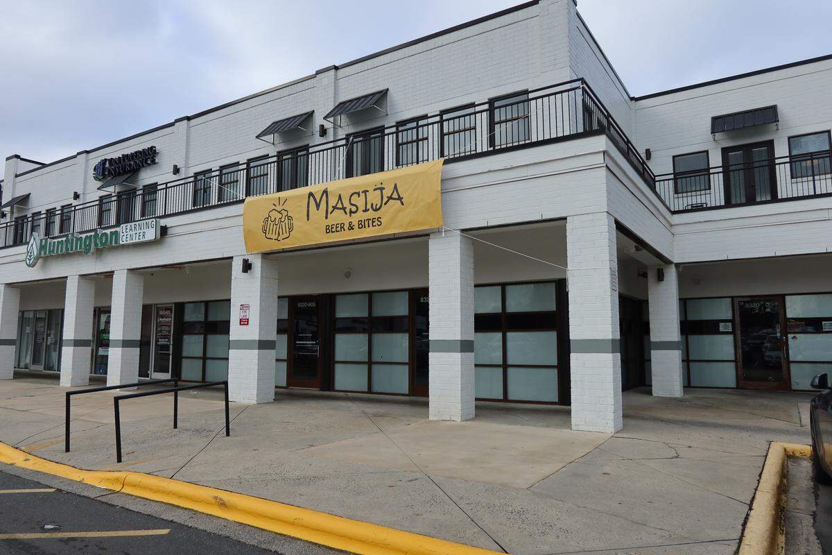The exterior of Masija Beer & Bites is shown, featuring a yellow banner over the entrance of a modern two-story white brick building. The storefront is located in a quiet shopping center with a Huntington Learning Center visible to the left.