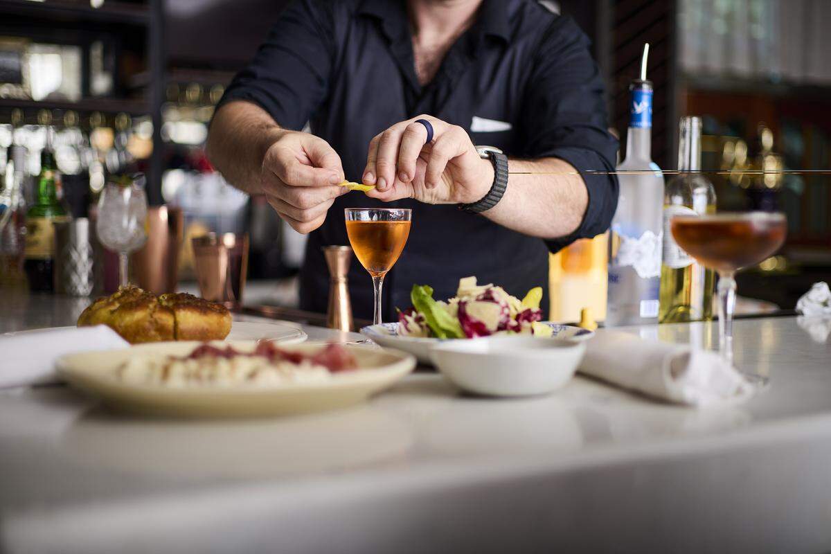 A bartender in a dark shirt is captured in a shallow-focus shot, meticulously twisting a lemon peel over a golden-colored cocktail. The bar counter in the foreground is lined with plates of appetizers, including focaccia and a meat and cheese platter. The background is softly blurred, highlighting the craft of the drink preparation within a dimly lit, sophisticated bar environment.