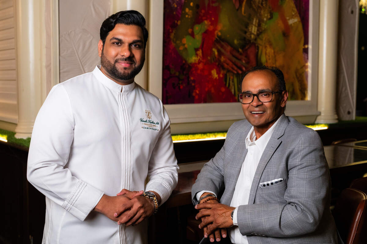 Two men are posing together indoors while looking directly toward the camera with pleasant smiles. The man on the left stands in a crisp white, high-collared chef’s coat with embroidered text, while the man on the right sits wearing a gray blazer over a white shirt and glasses. A colorful, abstract painting hangs behind them in the dimly lit, upscale venue.