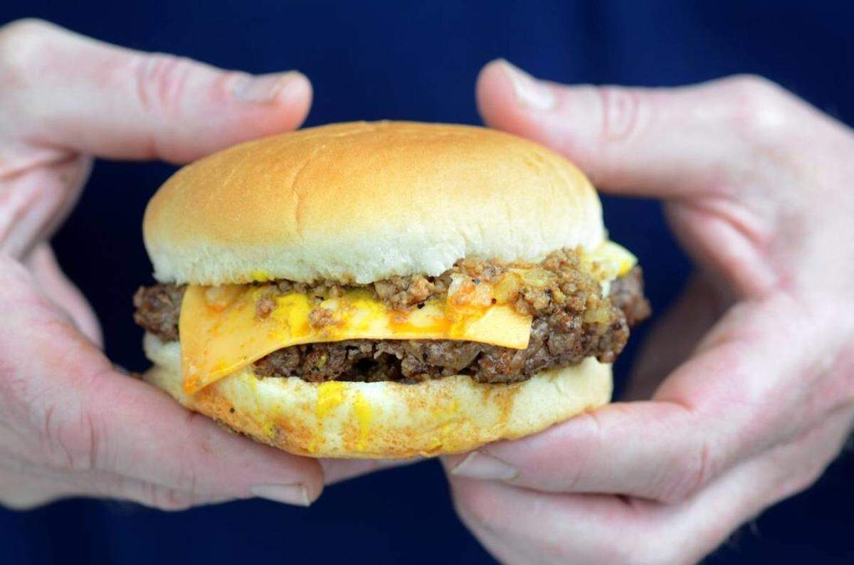 A pair of hands gently holds a small, classic cheeseburger against a blurred blue background. The burger features a soft, light-brown bun, a dark, well-done patty, a slice of melted yellow American cheese, a dollop of yellow mustard, and a generous layer of what appears to be minced onions or a chili-like topping on the patty. The bottom bun has some visible drippings.