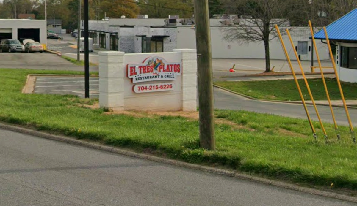 A low-angle, outdoor shot shows a white stone monument sign for “El Tres Platos Restaurant & Grill” situated on a grassy roadside. The sign features red lettering and a local phone number below a colorful logo. In the background, a parking lot and several commercial buildings are visible under a clear sky.