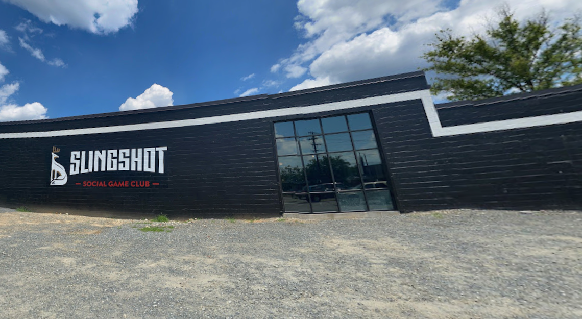 The exterior of a long, black brick building features a large white “Slingshot Social Game Club” logo with a stylized deer icon. A tall, gridded glass window reflects the sky, and a gravel lot sits in the foreground under a bright blue sky with white clouds.