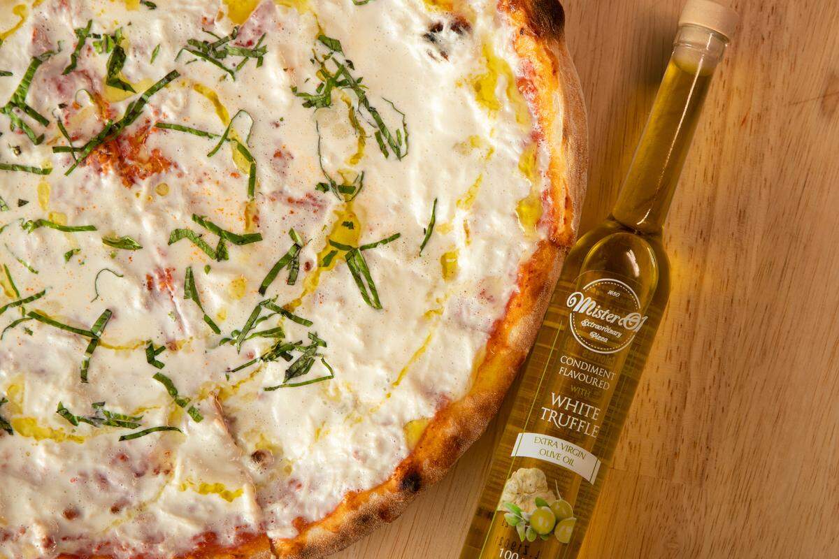 A close-up view of a white pizza topped with melted cheese and fresh basil sits next to a glass bottle of Mister O1 white truffle oil.