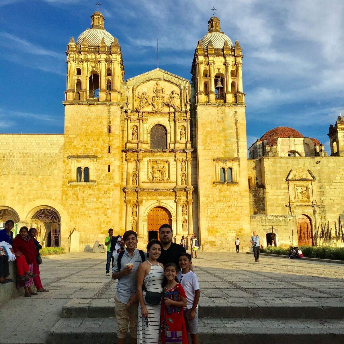 A golden-lit stone cathedral with two domed bell towers stands against a dramatic blue-gray sky. In the foreground, several people pose and smile while other visitors gather in the plaza around them. The scene feels like a busy, late-afternoon moment at a historic landmark.