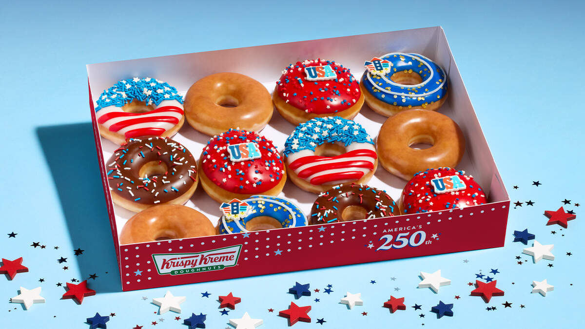An open Krispy Kreme doughnut box sits against a light blue background, filled with a dozen assorted doughnuts, including classic glazed, chocolate-iced with patriotic sprinkles, and red, white, and blue Fourth of July themed designs. Several doughnuts feature festive toppings like “USA” candy pieces, blue frosting with white star sprinkles, and red and white stripes. The front of the red box is decorated with white polka dots, the Krispy Kreme logo, and text celebrating “America’s 250th,” surrounded by small star confetti scattered on the surface below.