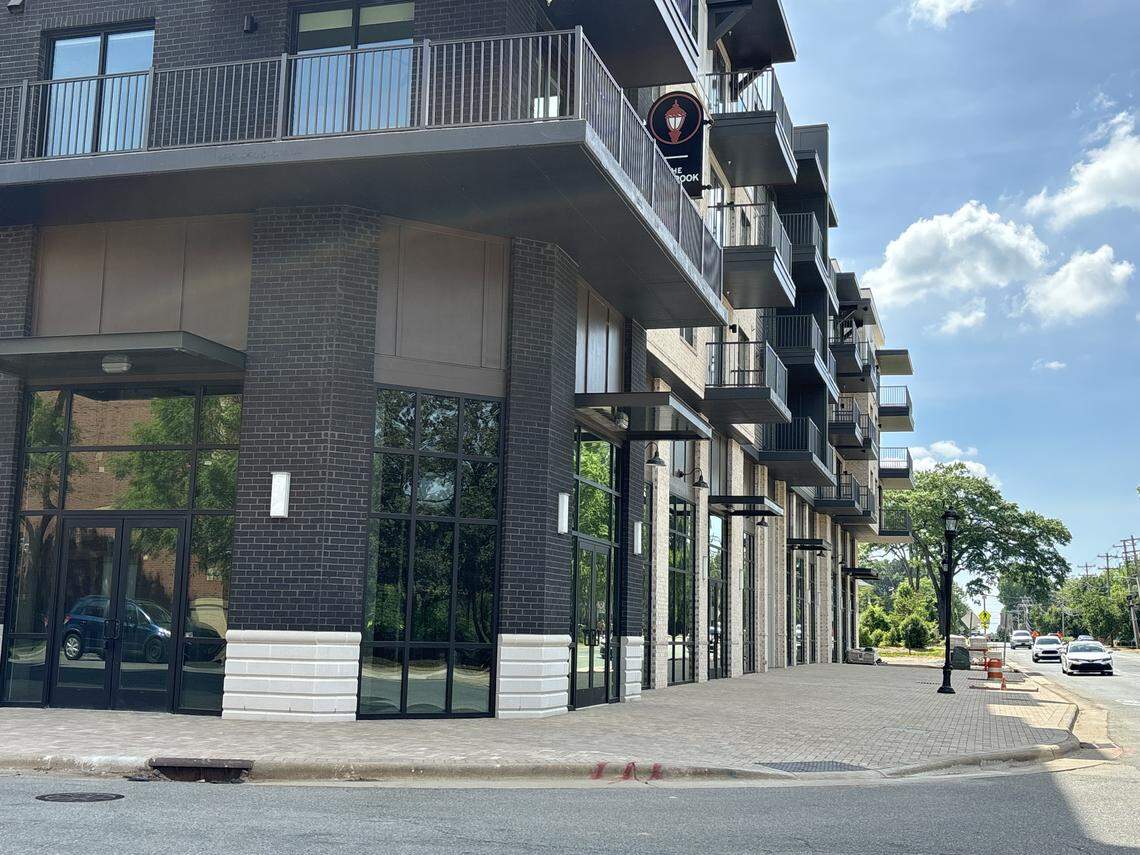 This low-angle exterior photo shows a modern apartment or mixed-use building with black brick, white stone accents, and several floors of dark metal balconies. The ground level features large storefront windows along a wide brick-paved sidewalk that stretches into the distance.