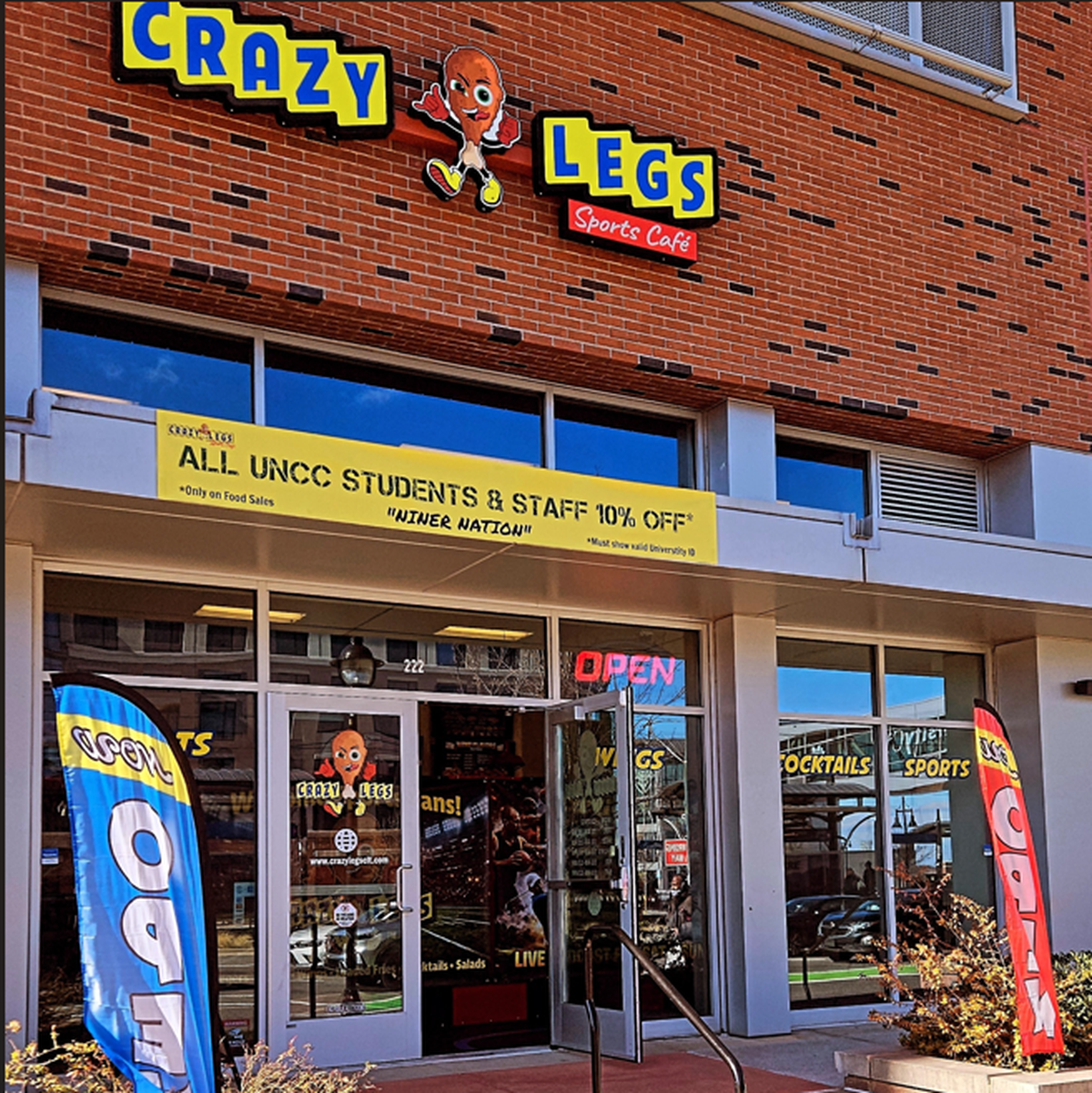 The exterior of Crazy Legs Sports Café shows a brick building with a bright yellow banner offering a 10% discount to UNCC students and staff. Colorful “Open” flags stand on the sidewalk near the glass entrance doors.