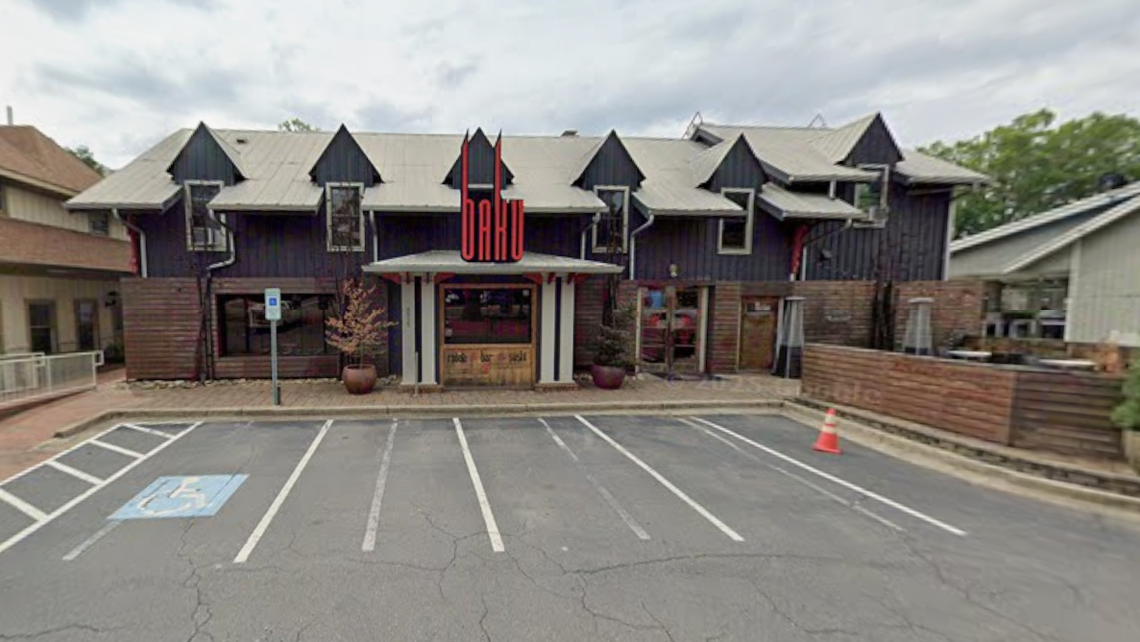 The front facade of a restaurant named Baku features dark siding, a gabled roofline with multiple dormer windows, and a prominent red vertical sign. The building is viewed from an asphalt parking lot that includes a designated accessible parking space.
