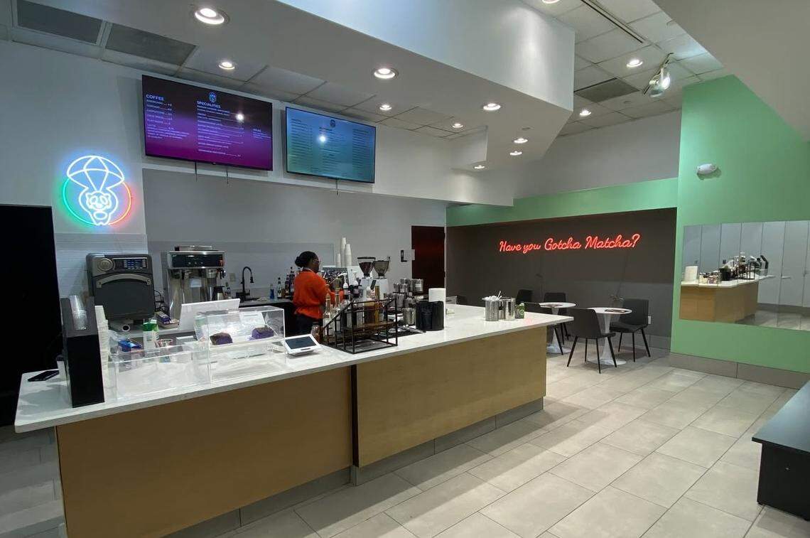 The interior of Gotcha Matcha features a clean, modern aesthetic with a long white service counter and a glowing neon panda logo. A red neon sign on the back wall reads “Have you Gotcha Matcha?” above a small seating area with black chairs and white cafe tables.