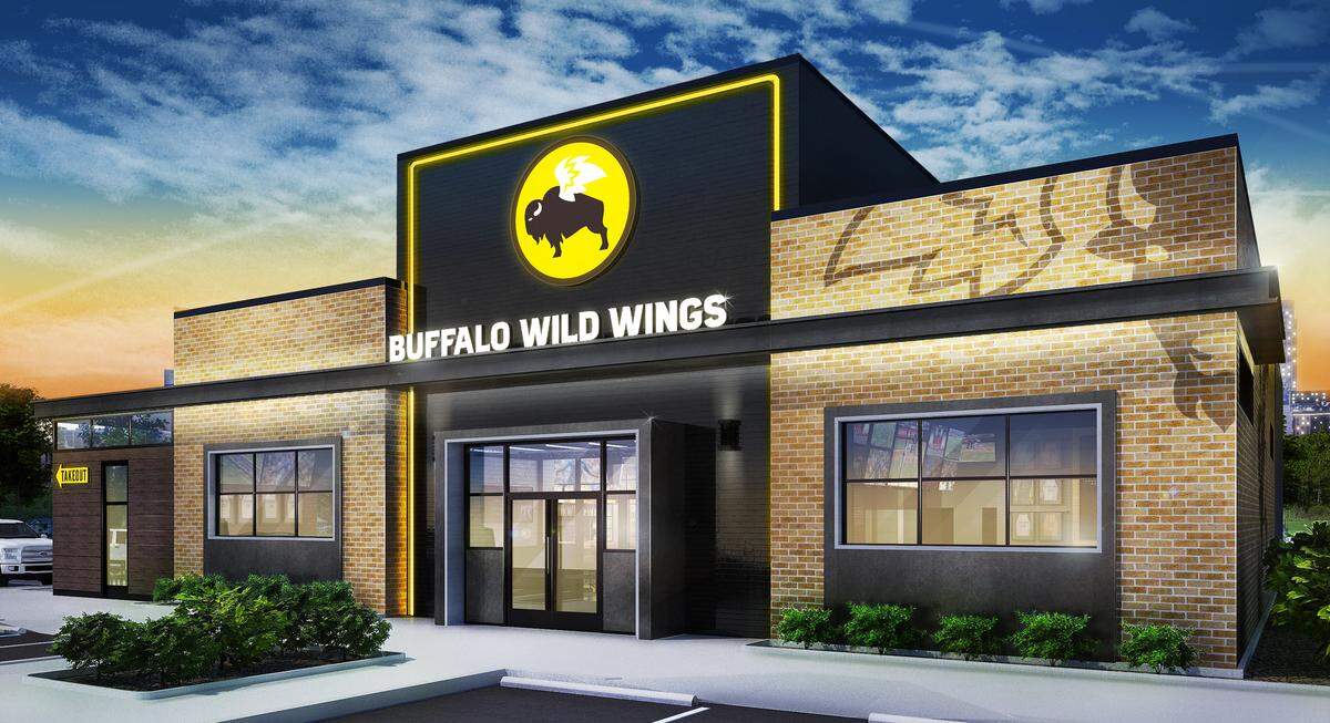 The exterior of a Buffalo Wild Wings building displays a sleek, modern facade with a mix of light brick and dark textured paneling. The iconic yellow and black buffalo logo is prominently centered above the glass entrance, illuminated by a glowing yellow neon border against the sunset sky.