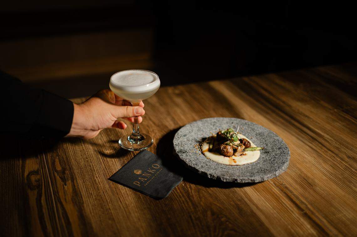 A person in a dark suit holds up a coupe glass containing a Pisco Sour cocktail with a white foam topping. Next to the hand is a small, dark napkin with the PANKO Peruvian Fusion logo and a stone-like plate holding a small plate of Starter Loin Bonbons.