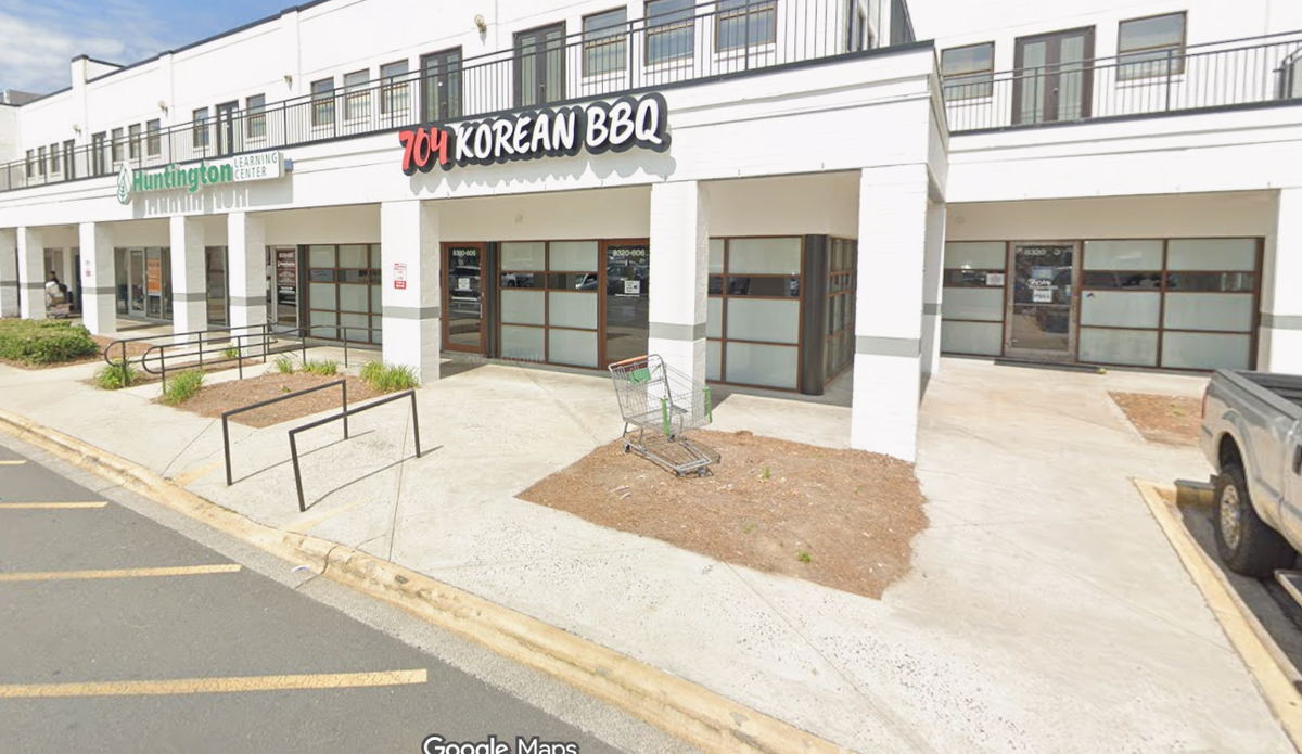 The exterior of the 704 Korean BBQ restaurant, which has a red and white sign. The restaurant is located in a white brick strip mall next to a Huntington Learning Center. The image also shows a sidewalk, some planted dirt patches, and a metal shopping cart resting in front of the main entrance.
